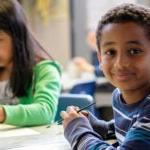 Two students sit at a table in a classroom; one student smiles for a photo